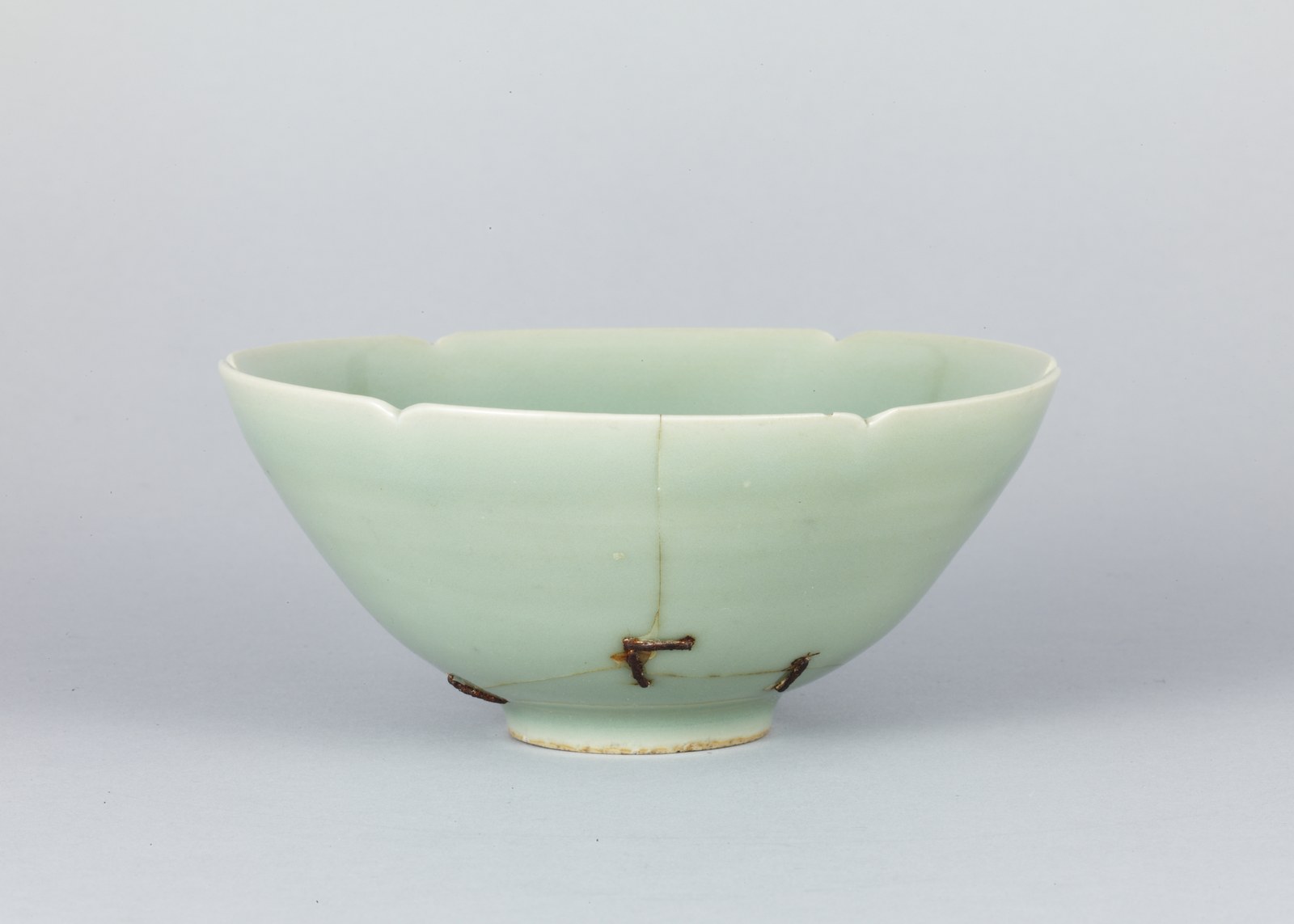 Pale green celadon tea bowl with a scalloped rim; a crack runs down its side and three metal staples hold it together near the foot
