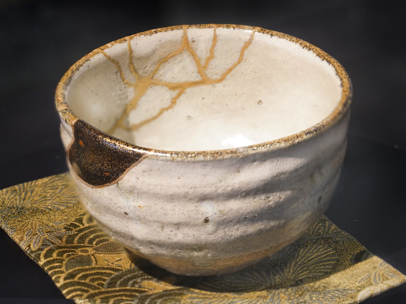 Cream-glazed Karatsu-ware tea bowl on a gold brocade cloth, with branching gold lines inside and a dark, gold-flecked patch filling a chip in the rim