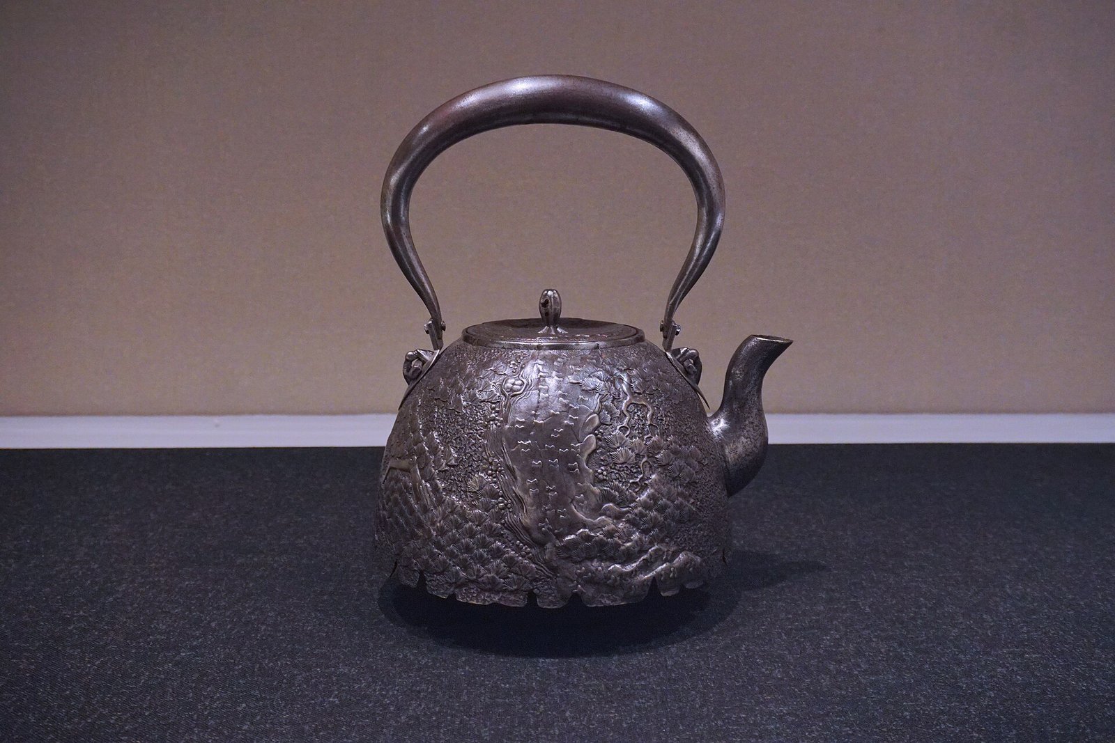 An old iron kettle with a raised pine-tree design on its body, a curved spout and an arched iron handle, displayed on a dark base against a plain wall.