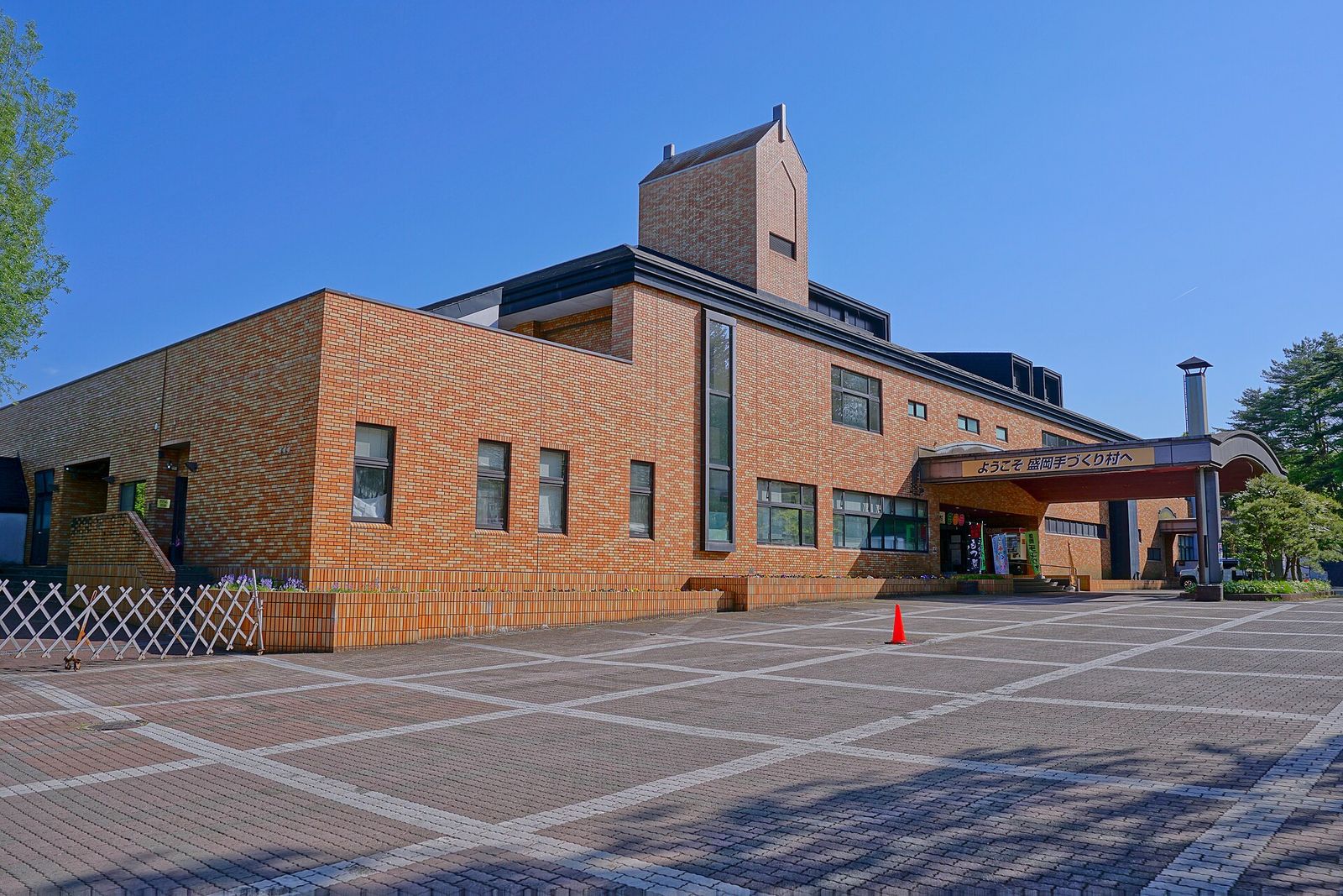 A brick building with a covered entrance and a welcome sign reading Morioka Tezukuri Mura in Japanese, behind a wide paved square under a blue sky.